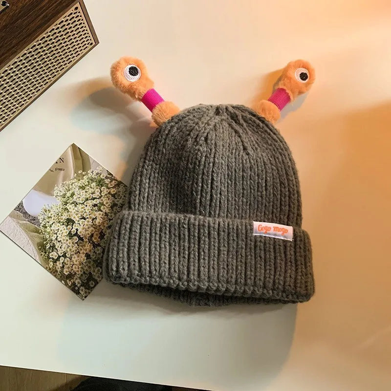Gray knit beanie with a small plush toy on top, placed on a surface with a blurred background.